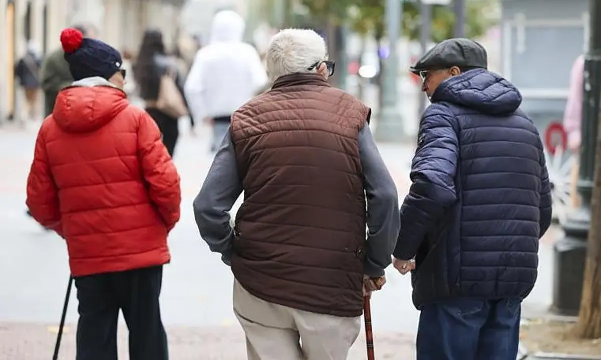 Tres personas mayores caminan por la calle, una con bastón, reflexionando sobre la **pensión mínima**.