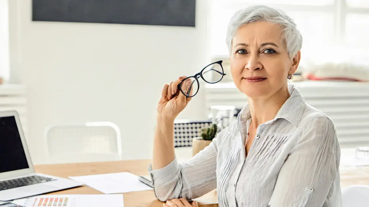 Mujer mayor con gafas, reflexionando sobre los tipos de jubilación. En su escritorio hay un portátil y documentos.