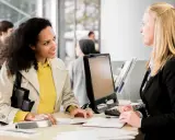 Mujer sonríe a empleada de banco mientras tramita su cuenta BBVA con pasaporte.