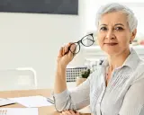 Mujer mayor con gafas, reflexionando sobre los tipos de jubilación. En su escritorio hay un portátil y documentos.