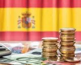 Monedas y billetes sobre una mesa, con la bandera de España de fondo. Una calculadora a la izquierda. Reflexión sobre qué son los tributos.