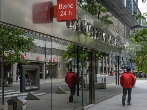 Un hombre con chaqueta roja camina por la acera frente a un edificio de **bpa finanzas** con un letrero de "Banc 24 h".