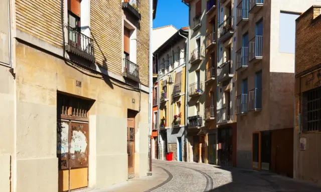 Calle adoquinada con edificios coloridos y balcones. Un cartel de "haya real estate" podría estar en uno de estos locales.