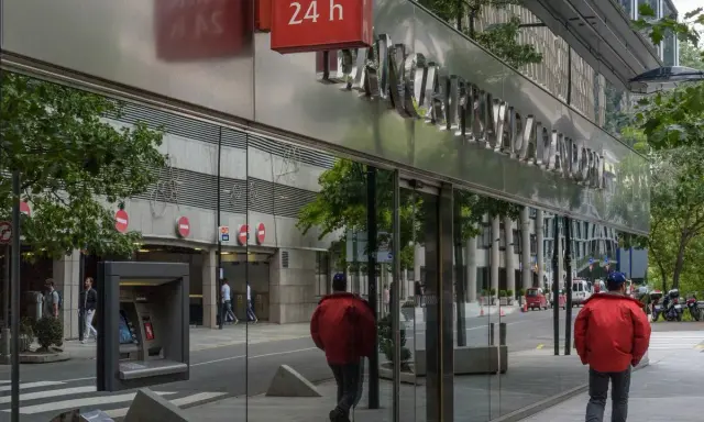 Un hombre con chaqueta roja camina por la acera frente a un edificio de **bpa finanzas** con un letrero de "Banc 24 h".