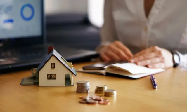 Maqueta de casa, monedas y portátil con gráfico. Persona anotando en libreta. Analizando el precio vivienda.