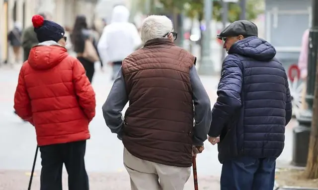 Tres personas mayores caminan por la calle, una con bastón, reflexionando sobre la **pensión mínima**.