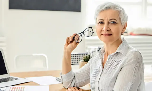 Mujer mayor con gafas, reflexionando sobre los tipos de jubilación. En su escritorio hay un portátil y documentos.