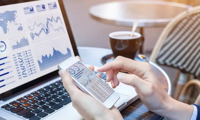 Manos revisando gráficos financieros en un teléfono móvil y portátil, gestionando el bbva net cash.