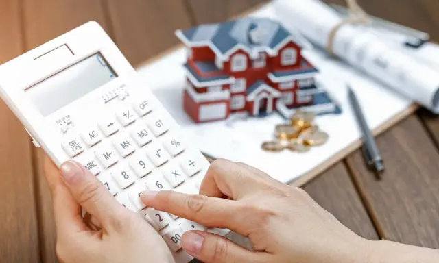 Calculando la casilla 435 renta: una mano usa una calculadora blanca junto a una maqueta de casa, monedas y planos.