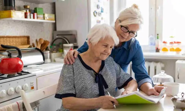 Dos mujeres mayores revisan documentos en la cocina. Una apoya a la otra, quizás tramitando la pensión de orfandad mayores de 30 años.