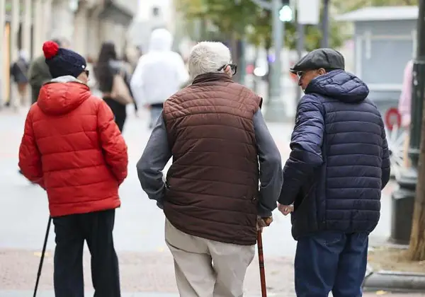 Tres personas mayores caminan por la calle, una con bastón, reflexionando sobre la **pensión mínima**.